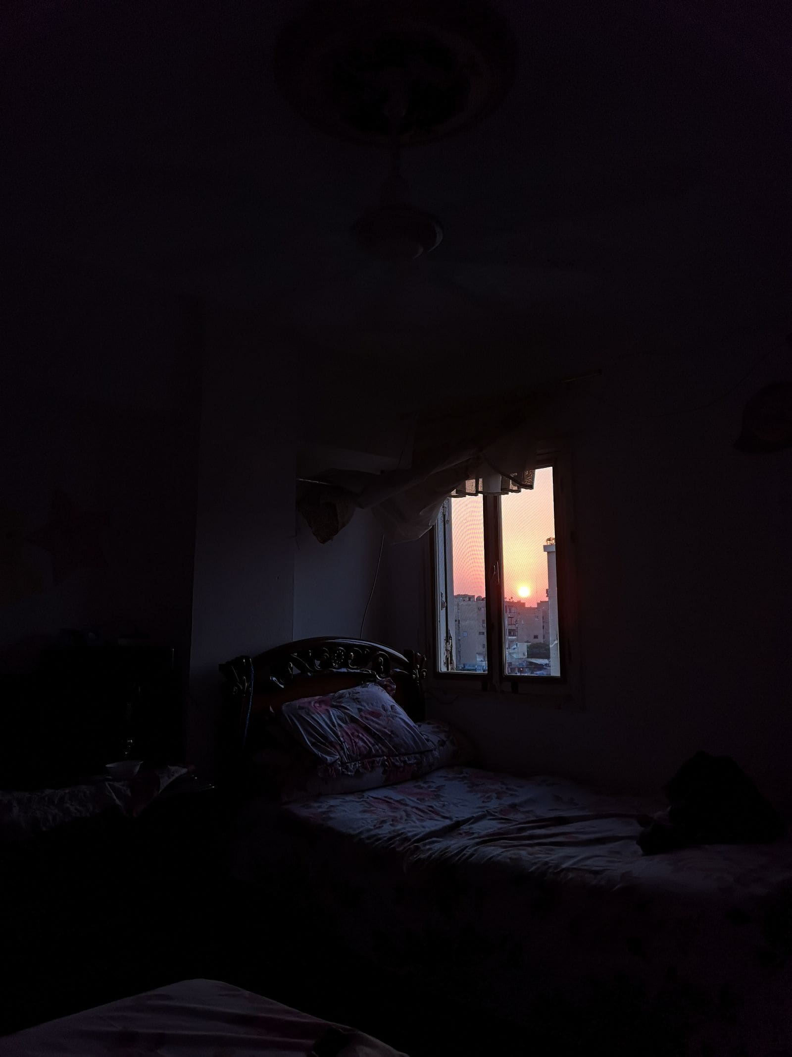 Bed in a dark bedroom beside a glass window with a sunset view
