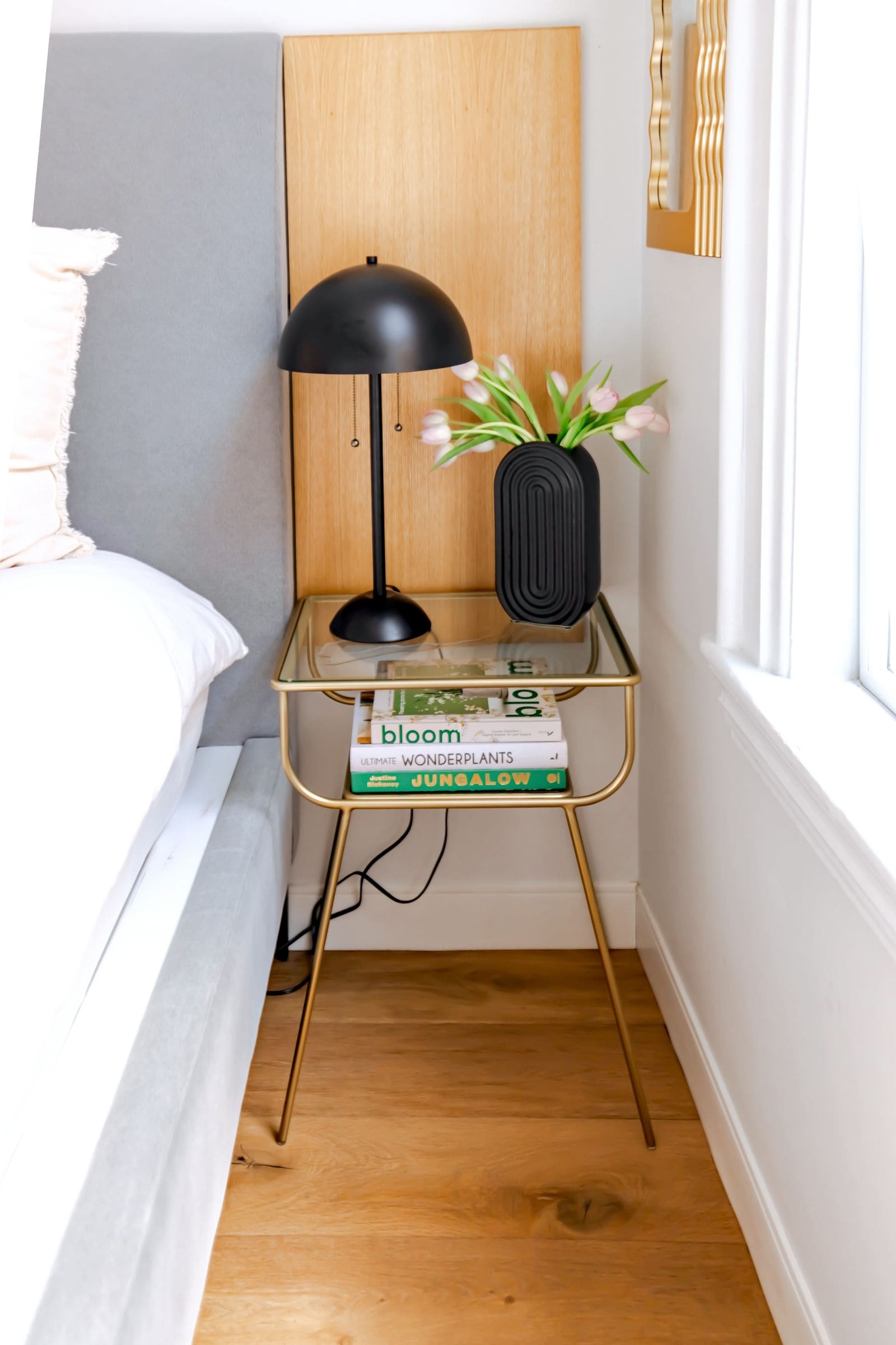 Bedside table with a lamp, books and a vase of flowers next to a made bed